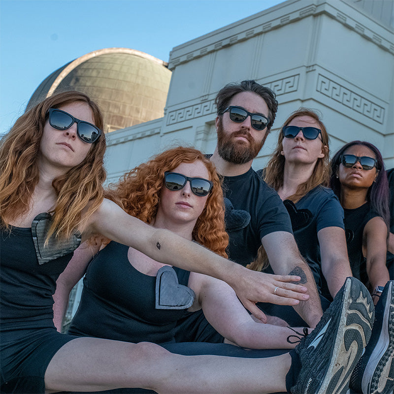 Five redheads in black sunglasses on an outdoor staircase stare into the camera while stretching their left leg on a railing.