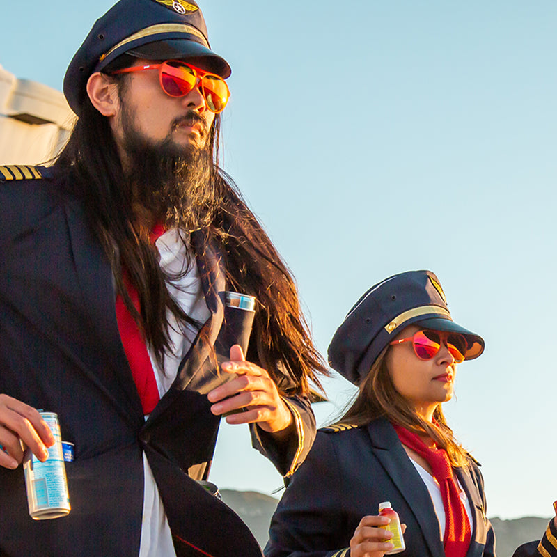 Two disheveled pilots wearing red aviator sunglasses with red lenses exit an airplane, energy drinks shoved in their pockets.
