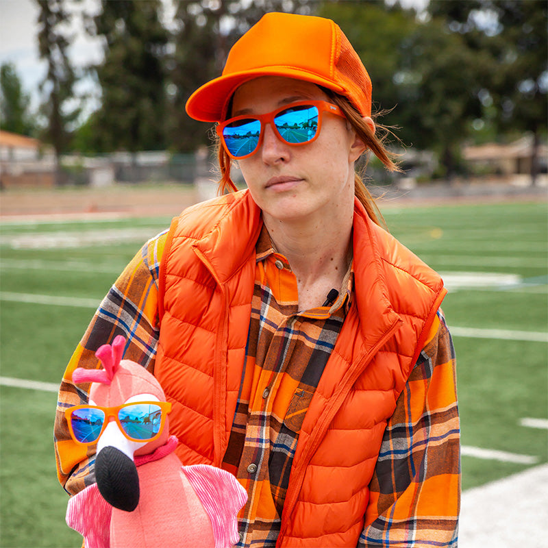 A woman in a bright orange outfit holds a stuffed pink flamingo, both of them wearing orange sunglasses with blue lenses.