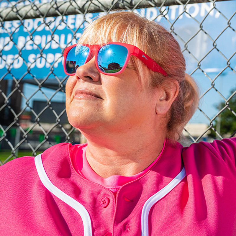 A woman in a hot pink baseball uniform and hot pink wide-fit sunglasses with blue lenses leans against a fence in a sunbeam.