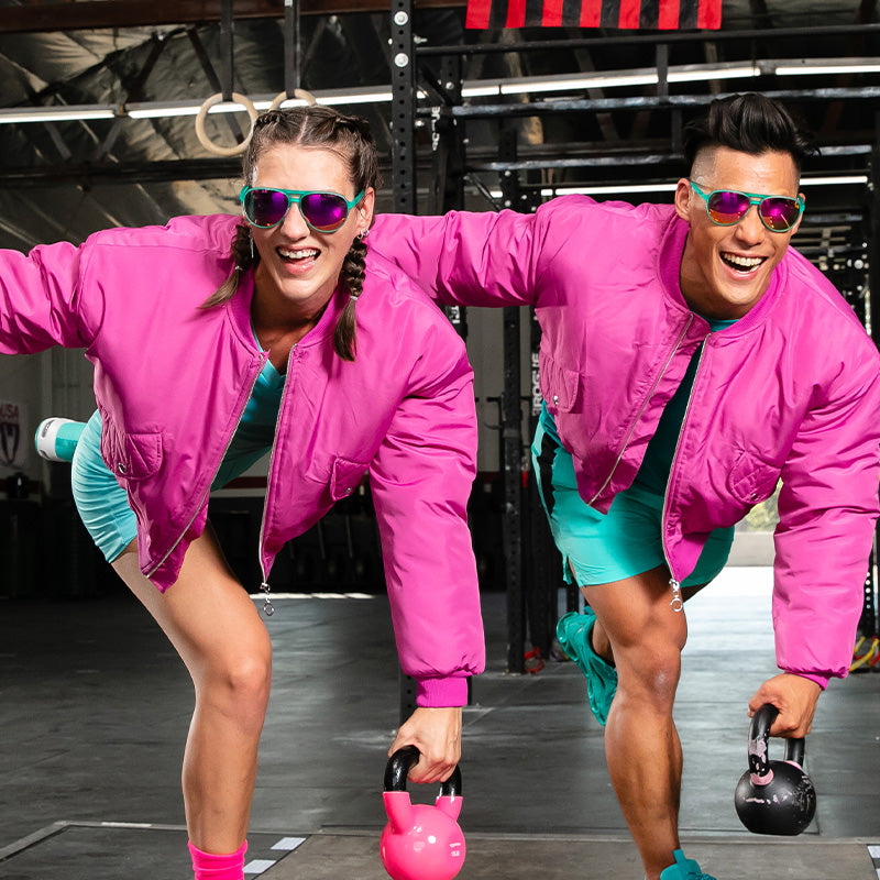 A smiling man and woman in pink bomber jackets and teal aviators with pink reflective lenses do a kettlebell workout.
