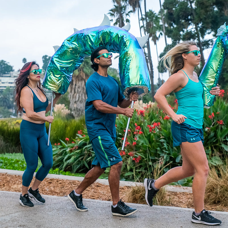 Three runners wearing teal sunglasses with teal lenses hold sections of a giant Loch Ness monster puppet as they jog.