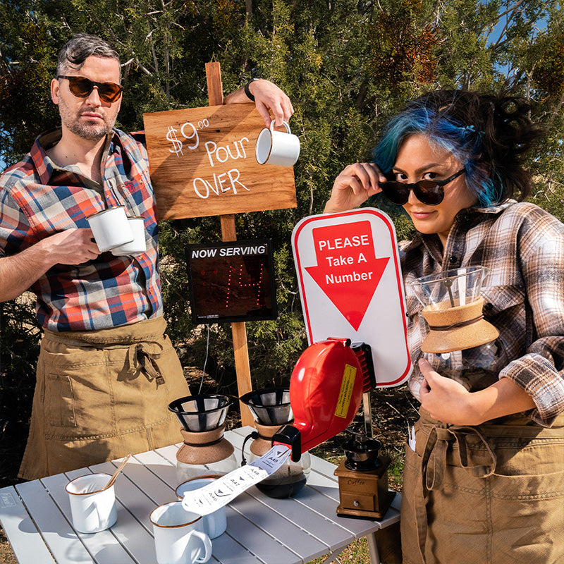 Two hipster baristas wearing round brown sunglasses with brown lenses pose at an indulgently artisanal pour-over coffee stand.