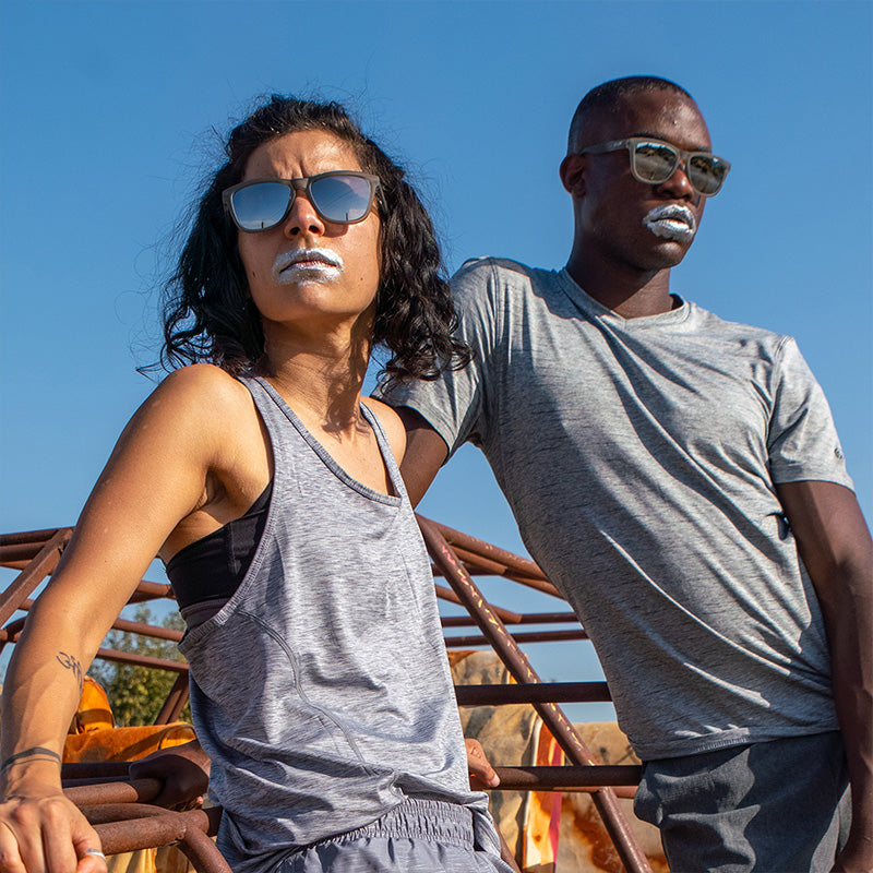 A man and woman wearing gray sunglasses with gray lenses and silver paint on their mouths look off in the distance fiercely.