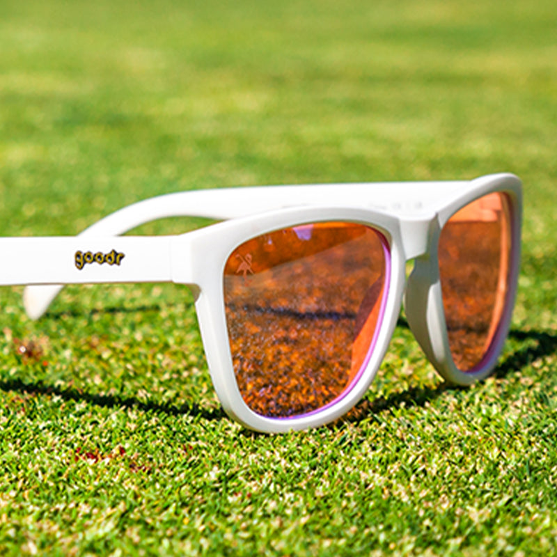 Three-quarter angle view of white sunglasses with rose-tinted lenses sitting on a blood-stained golf green.