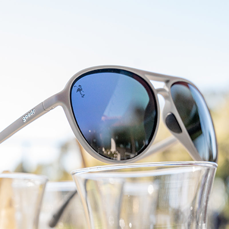 Three-quarter angle view of gray aviator sunglasses with black lenses perched atop a display of clear cocktail glasses.