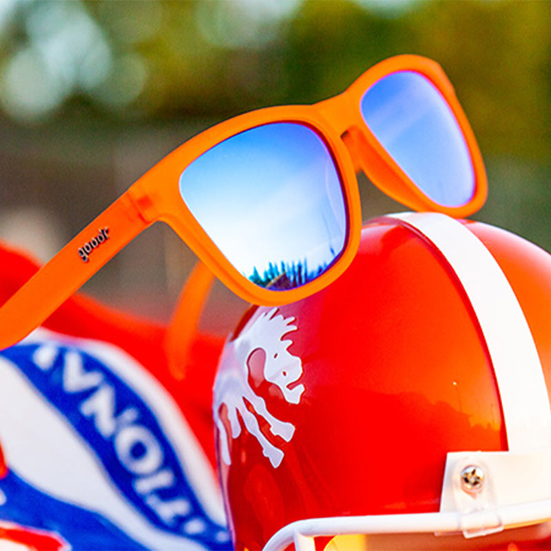 Three-quarter angle view of bright orange sunglasses with blue reflective lenses sitting on a bright orange football helmet.