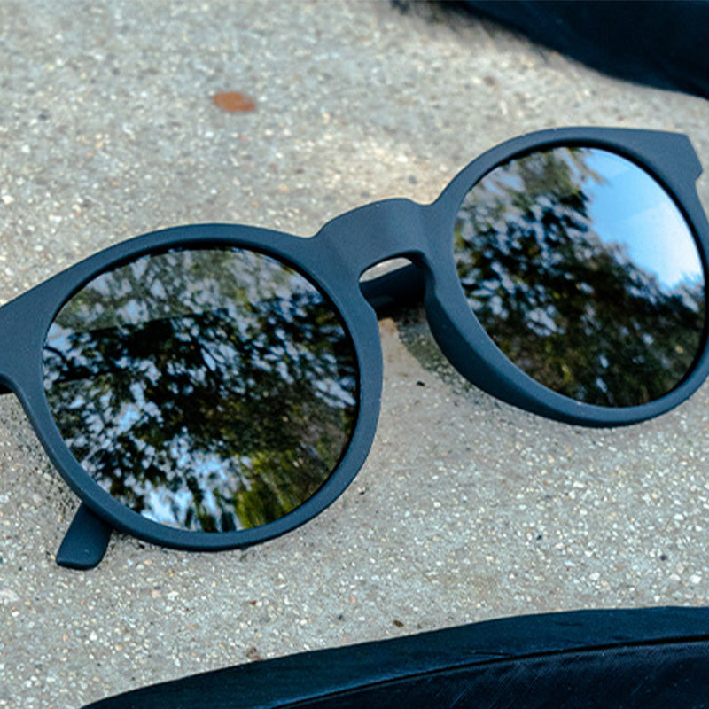 Three-quarter angle view of round black sunglasses with non-reflective black lenses sitting folded on concrete.