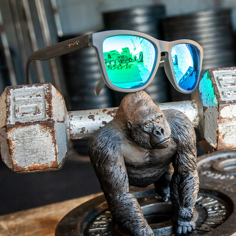 Three-quarter angle view of gray sunglasses with green lenses sitting atop a dumbbell, beside a gorilla action figure.