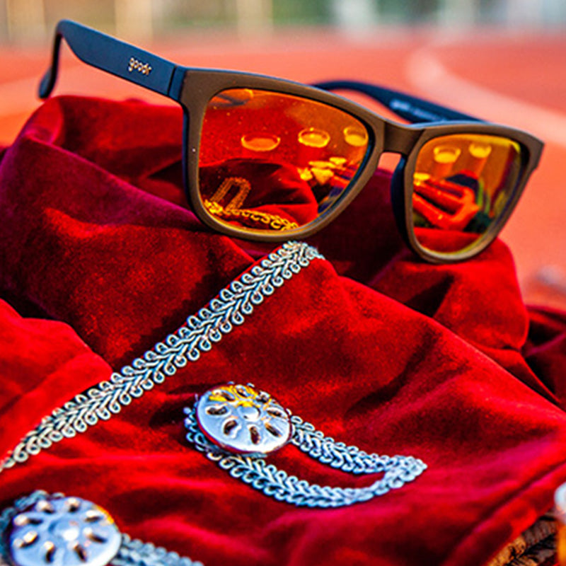 Three-quarter angle view of black sunglasses with amber reflective lenses sitting atop a red velvet coat on a running track.