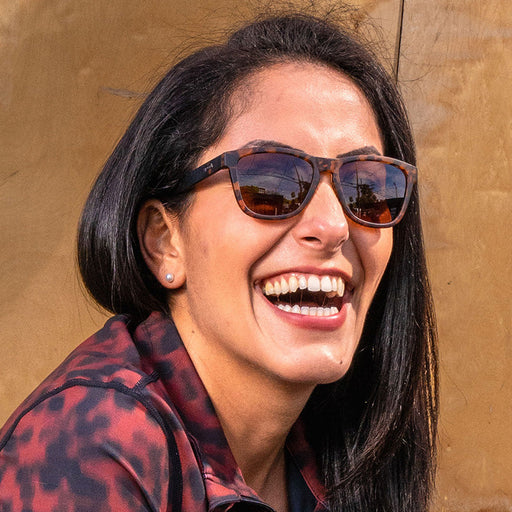 A woman with dark hair wearing brown tortoiseshell sunglasses with brown lenses laughs in the sunshine.