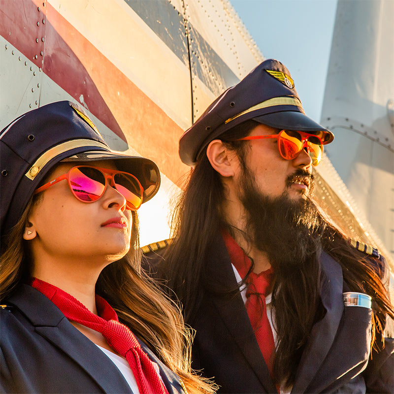 Two disheveled pilots wearing red aviator sunglasses with red lenses lean against an airplane, looking into the sunset.