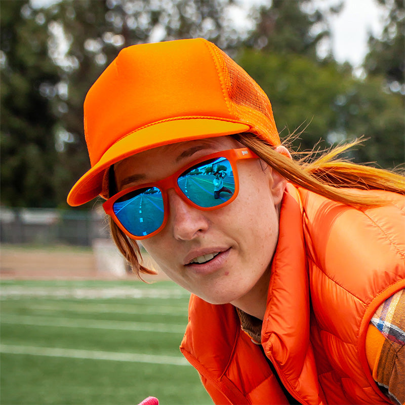 A woman in a bright orange outfit stands on a football field wearing bright orange sunglasses with blue mirrored lenses.