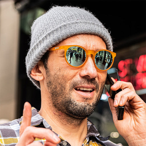 A hipster man in a gray beanie and polarised round orange sunglasses looks off to the distance while talking on a flip phone.