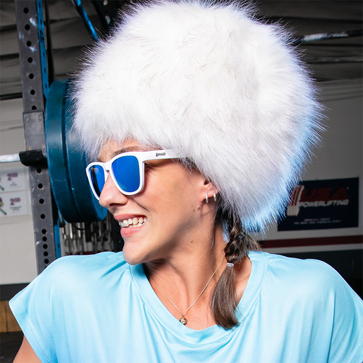 A woman in a white furry hat and white sunglasses with blue mirrored lenses looks to the side, a weight bench behind her.