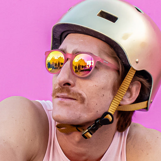 A stylish man in a gold bike helmet wears round pink sunglasses with pink lenses, looking off to the side.
