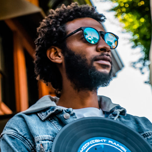 A hipster man wearing round black sunglasses with blue reflective lenses looks into the distance, holding a vinyl record.