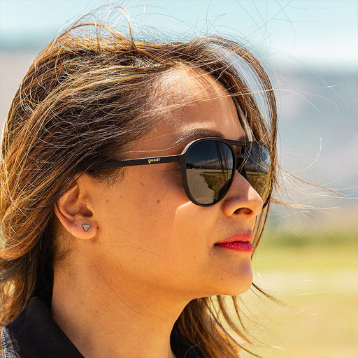 A woman in a black jumpsuit and black aviator sunglasses with black non-reflective lenses looks ahead fiercely.