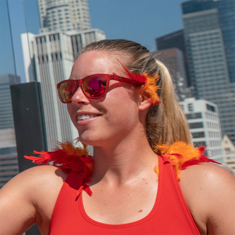 A woman wearing red sunglasses with red lenses with red and orange feather accents, a city skyline behind her.
