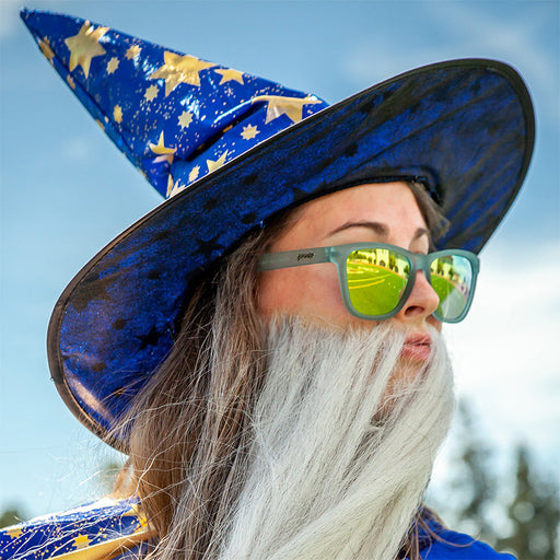 A woman in a wizard hat and big fake beard looks out, wearing translucent light blue sunglasses with gold reflective lenses.